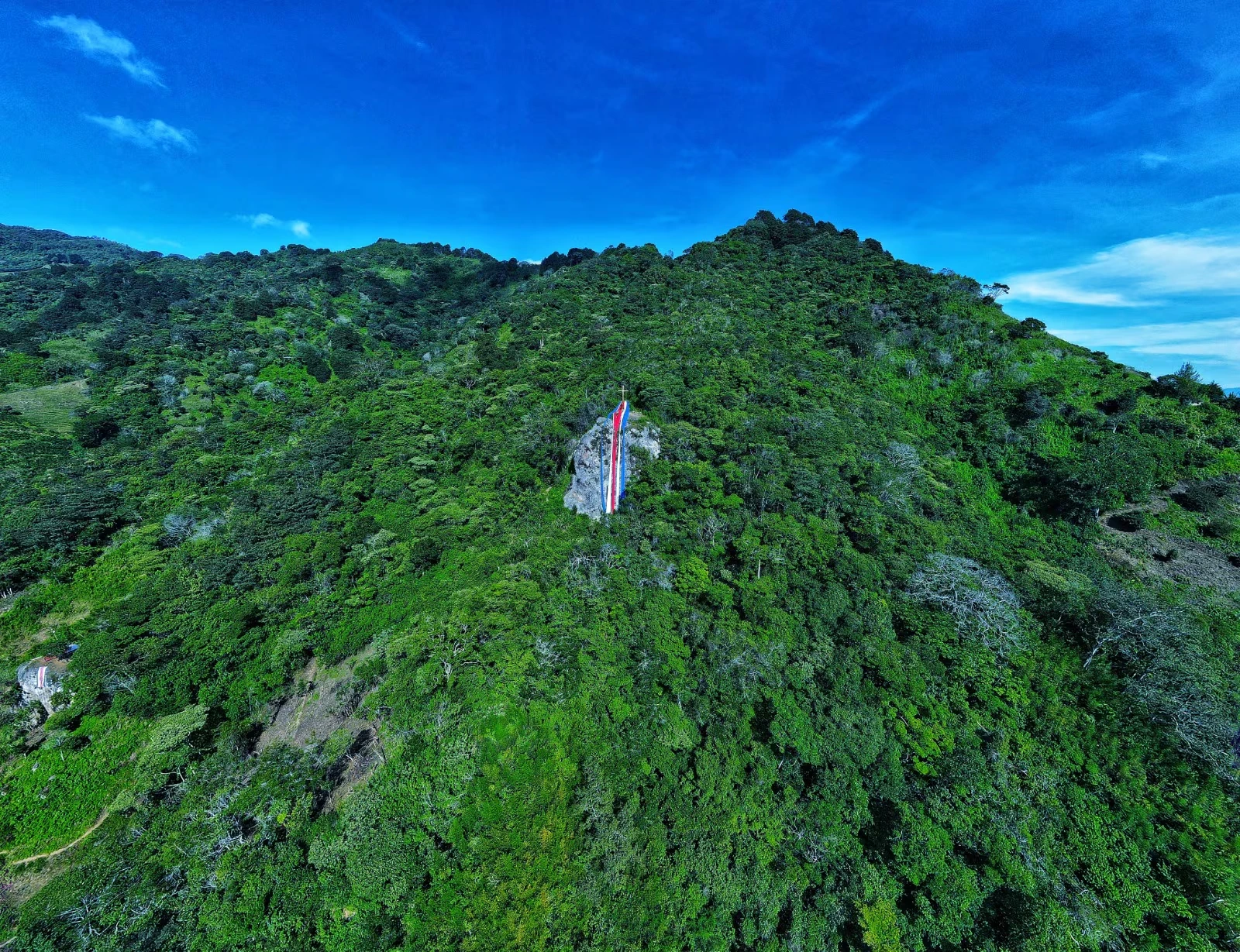 La Piedra de Aserrí sobresale entre las laderas verdes, con su cruz y una bandera de Costa Rica.
